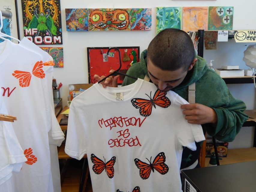 A person holds and closely examines a white t-shirt with red graffiti text “Migration Is Sacred” and orange monarch butterflies printed on it, inside an art studio.