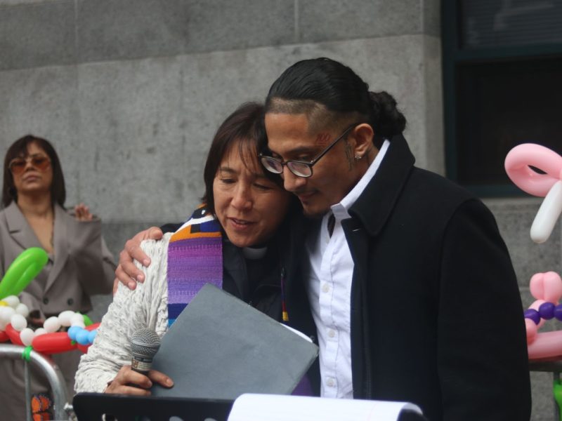 Two people stand close together, one with an arm around the other, holding papers. Balloons decorate the background, and another person stands behind them.