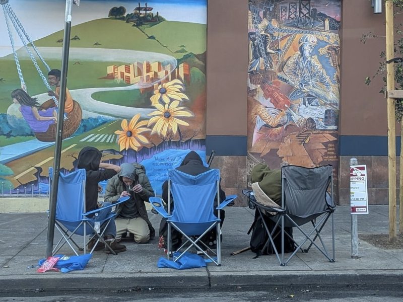 Four people sit in folding chairs on a sidewalk in front of a colorful mural, with scattered belongings nearby, as a marshall stands watchfully at the edge of the scene.