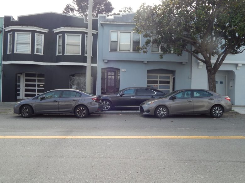 Three cars are parked in a row on a residential street in front of two attached houses, one painted dark gray and the other light blue.