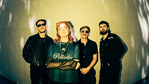 Four people stand in front of a light-colored wall, with shadows of leaves on the surface. The person in front wears a "Pelador" shirt; the others are in dark clothing and sunglasses.