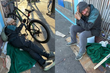 A man sits and lies on the sidewalk near a bicycle and a blanket, surrounded by personal belongings and makeshift bedding on a city street.