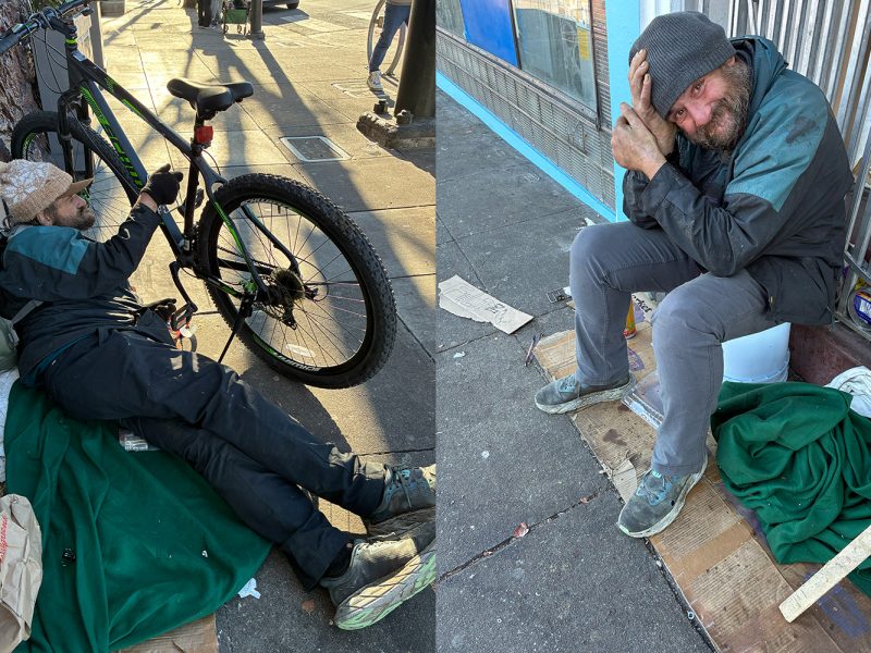 A man sits and lies on the sidewalk near a bicycle and a blanket, surrounded by personal belongings and makeshift bedding on a city street.