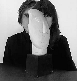 Two people in dark clothing sit behind a modern abstract sculpture, aligning their faces with the sculpture’s shape to create a visual blend of human and artwork.