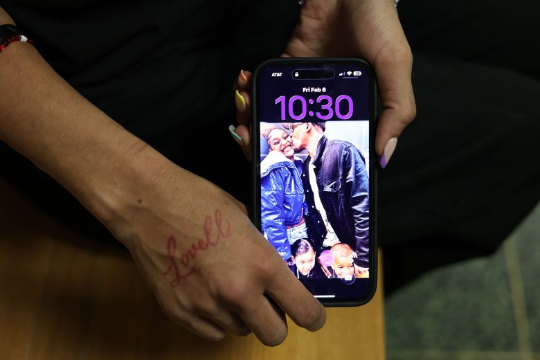 A person holds a phone displaying the time 10:30 and a lock screen photo of a man kissing a child, with two other children smiling. The person has "Lovell" written on their hand.