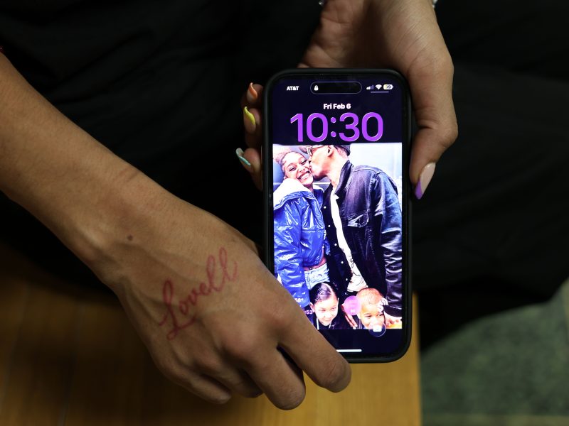 A person holds a phone displaying the time 10:30 and a lock screen photo of a man kissing a child, with two other children smiling. The person has "Lovell" written on their hand.