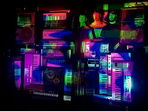 Three people stand behind a wall of electronic keyboards and synths, illuminated by colorful, multicolored lights and reflections.