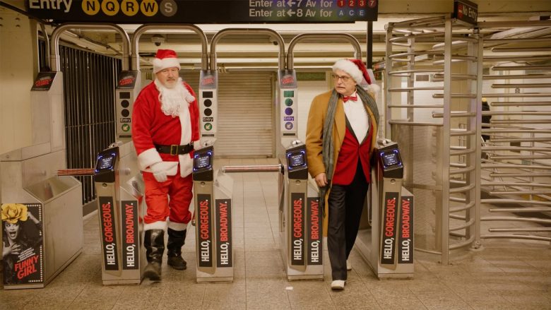 Two men, one dressed as Santa and one in a tan jacket with a Santa hat, stand next to turnstiles in a subway station. A "Funny Girl" poster is visible on the left.