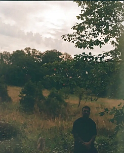 A person in dark clothing sits under a tree in a grassy field with trees in the background and a cloudy sky overhead.