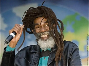 A man with long dreadlocks and a gray beard holds a microphone and smiles, standing in front of a backdrop with a globe design.