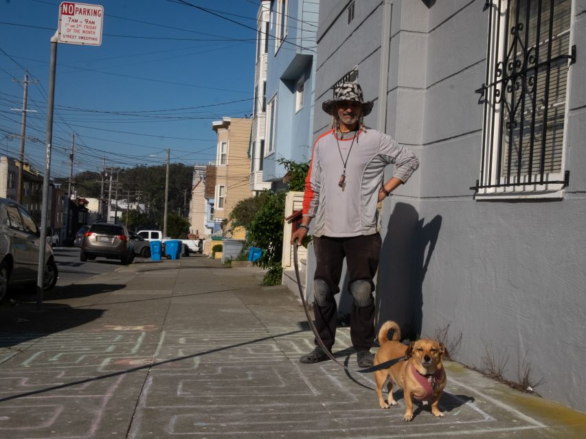 A person wearing a hat and sunglasses stands on a sidewalk holding a small dog on a leash; chalk drawings cover the pavement.