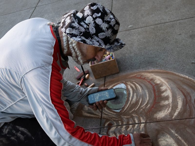 A person wearing a patterned hat and holding a phone creates chalk art on pavement, with pastel sticks and a box nearby.
