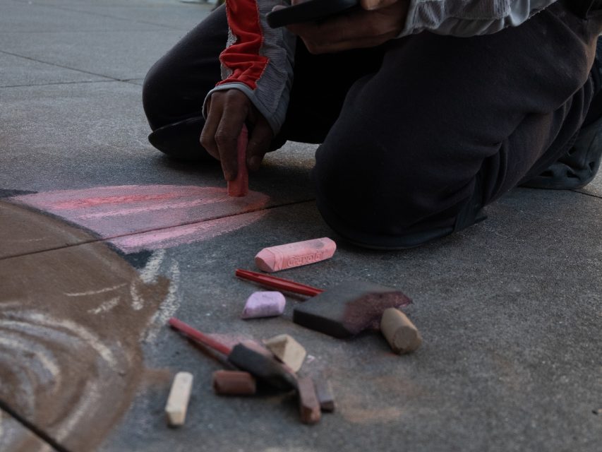 A person kneels on pavement drawing with large colored chalk pieces, holding a phone, with several chalk sticks scattered nearby.