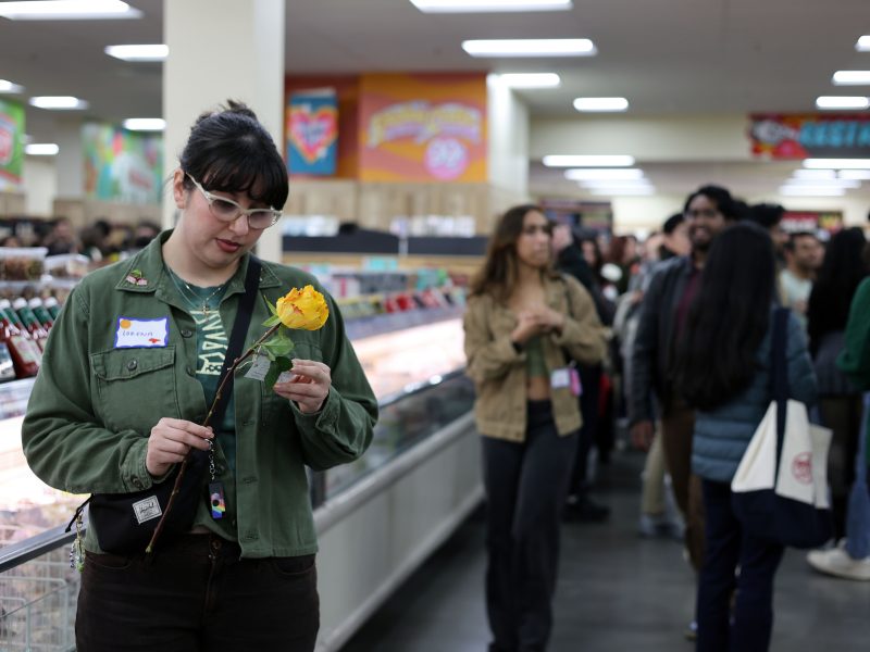 A person wearing glasses and a green jacket holds a yellow flower and looks at it in a crowded grocery store aisle.