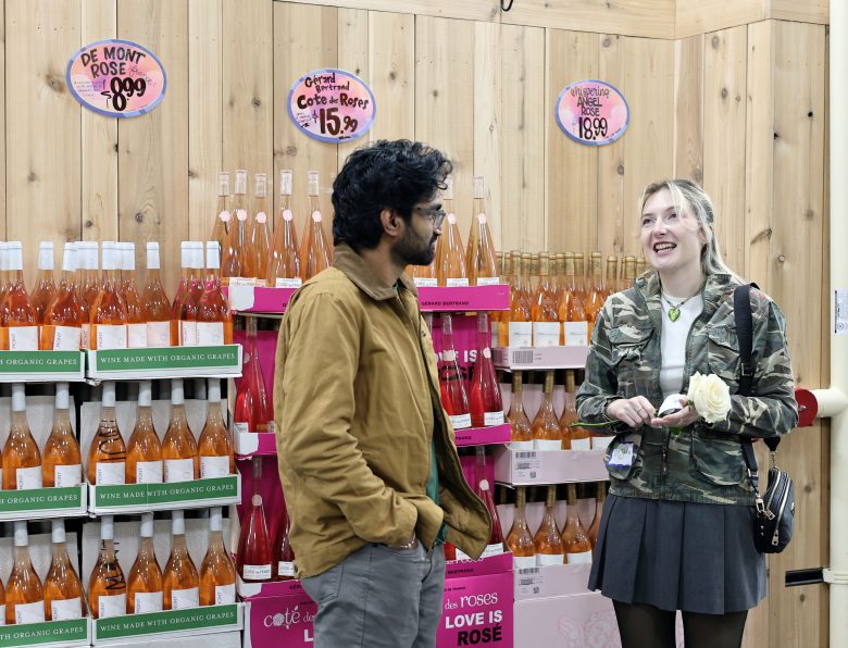 Two people stand and talk in front of shelves stocked with bottles of rose wine at a grocery store. Signs above display prices for different varieties.