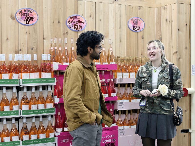 Two people stand and talk in front of shelves stocked with bottles of rose wine at a grocery store. Signs above display prices for different varieties.
