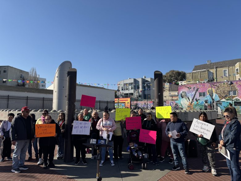 A group of people stand outdoors holding colorful protest signs in front of a mural and some buildings on a sunny day.
