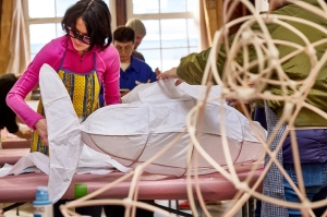 Several people work on constructing large animal-shaped sculptures using white fabric and wooden frames in a well-lit room.