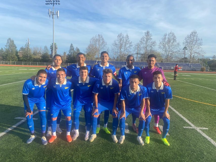 A soccer team of eleven players in blue uniforms poses together on a field, with trees and a running track in the background.