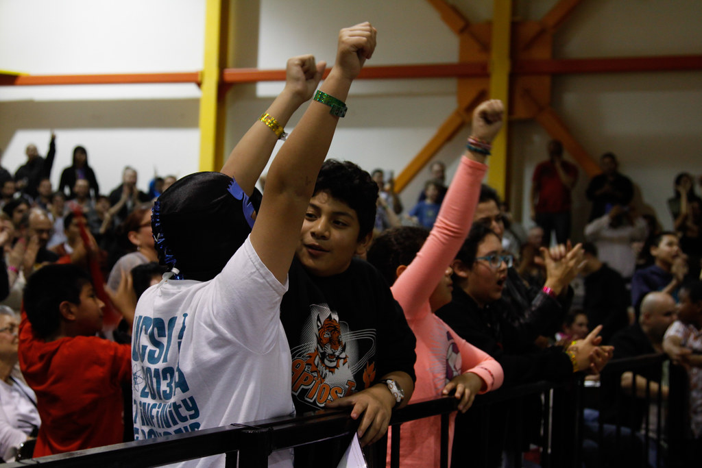 Children and adults cheer with raised fists behind a barrier at an indoor event, with a crowd visible in the background.
