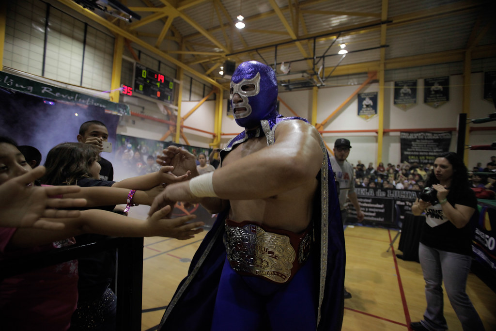 A masked wrestler wearing a blue cape and championship belt greets fans in a gymnasium during an event.