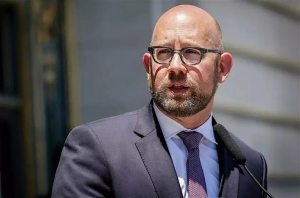 A man in a suit and tie speaks into a microphone outdoors.