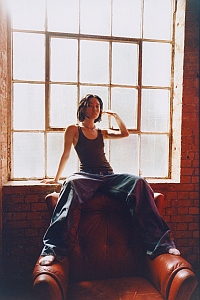 Person wearing a tank top and wide-leg jeans sits on the backrest of an armchair in front of a large industrial-style window with diffused light.