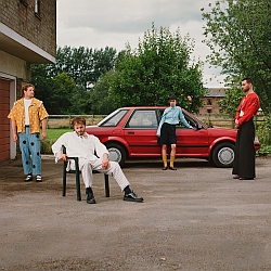 Four men in colorful, eclectic outfits pose near a red car parked outside, with trees and a building in the background. One man is seated on a plastic chair.