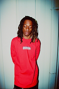 A person with long dreadlocks stands against a light-colored wall, wearing a red long-sleeve shirt with a rectangular design on the chest and looking downward.