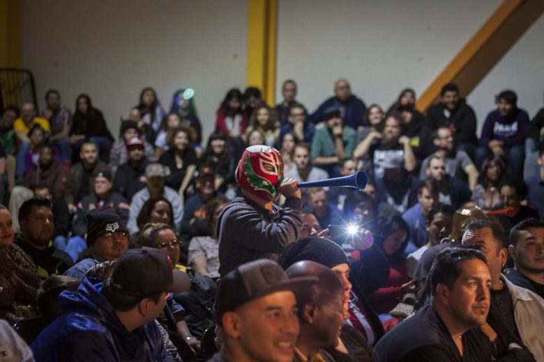 A person in a mask blows a vuvuzela in a crowded indoor audience while others watch an event.