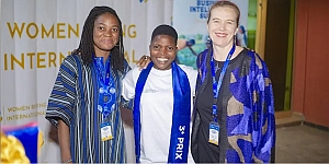 Three women stand together smiling at a formal event, one wearing a sash that reads "3e PRIX," with a banner in the background mentioning "WOMEN" and "INTERNATIONAL.