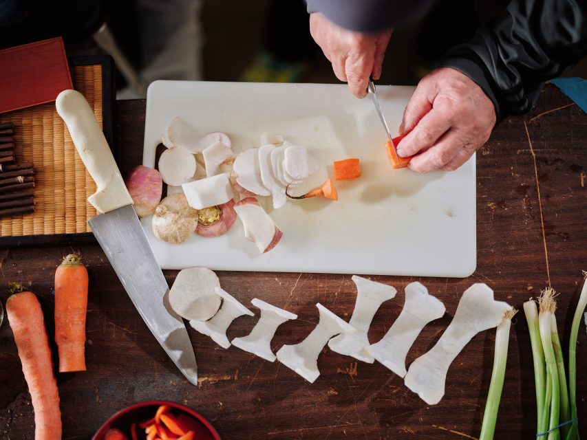 A person slices root vegetables on a white cutting board with a knife; carrots, daikon and green onions are visible on a wooden surface.