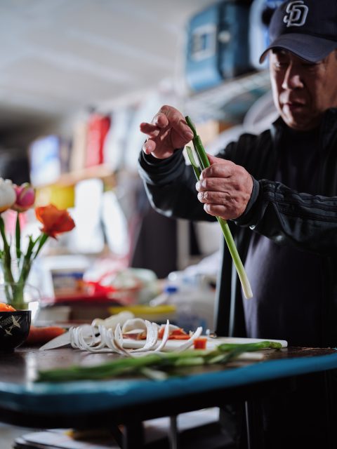 A man in a dark jacket prepares green onions at a kitchen counter, with sliced onions and flowers visible nearby.