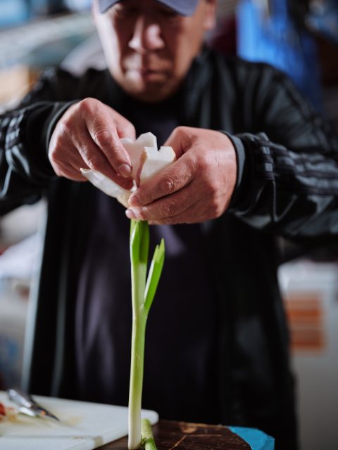 A person peels the outer layer off a green onion with their hands, with a cutting board visible in the foreground.