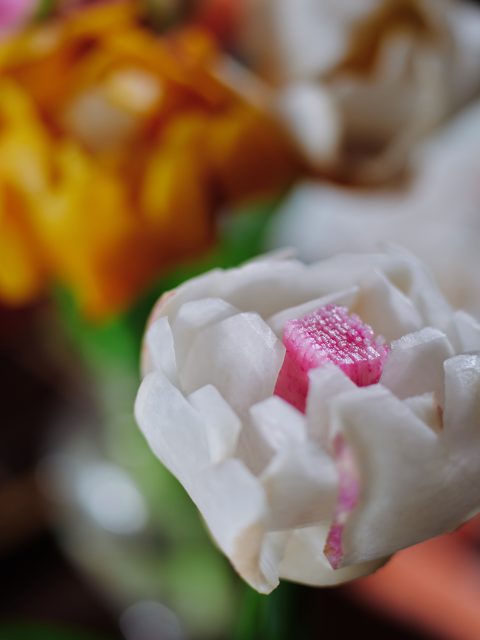 Close-up of a white and pink artificial flower, with blurred orange and white flowers in the background.