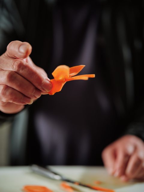 A person holds up a thin, curly slice of carrot with one hand, while other carrot slices and a peeler are visible on the table below.