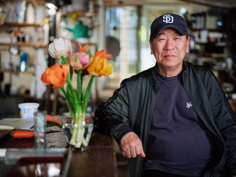A man wearing a dark jacket and a San Diego Padres cap sits at a table with colorful flowers in a vase in a cluttered, sunlit room.