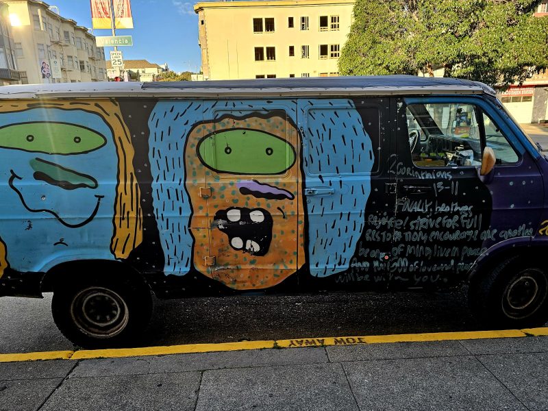 A van parked on the street features colorful cartoon faces and handwritten text painted on its side.