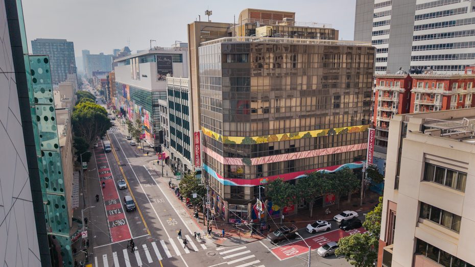 A city street with a glass office building on the corner, traffic lanes, crosswalks, trees, and several cars, surrounded by other tall buildings.