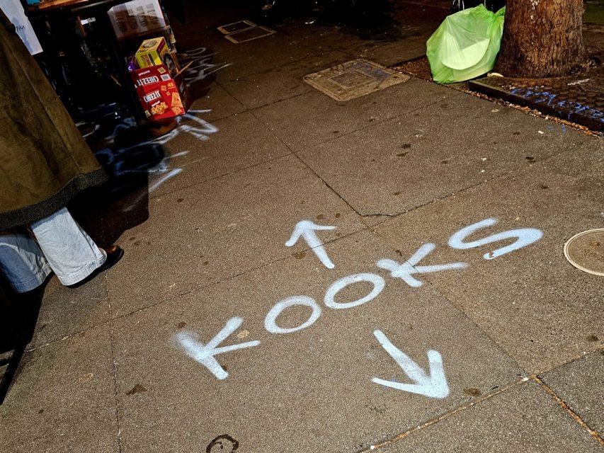 The word "KOOKS" and two arrows are spray-painted on a sidewalk at night, with boxes and a tree visible nearby.