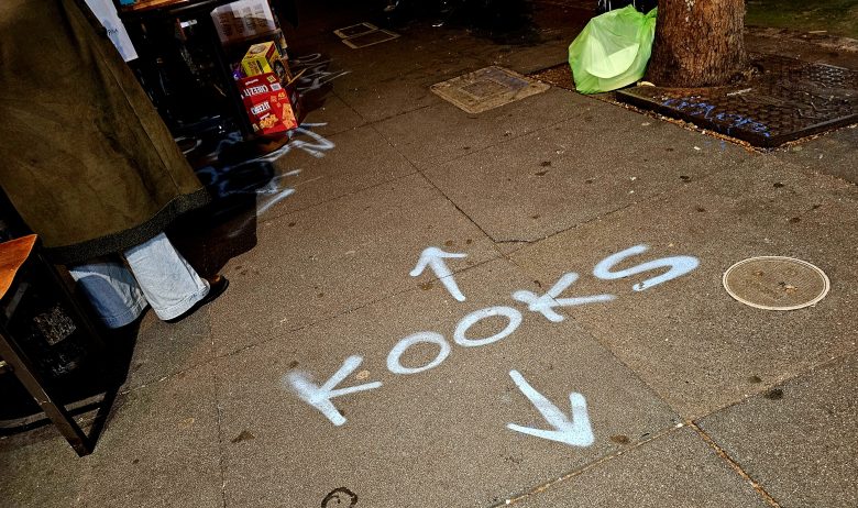Spray-painted arrows and the word "KOOKS" on a city sidewalk; nearby are boxes, a green bag, a tree, and part of a person's legs visible.