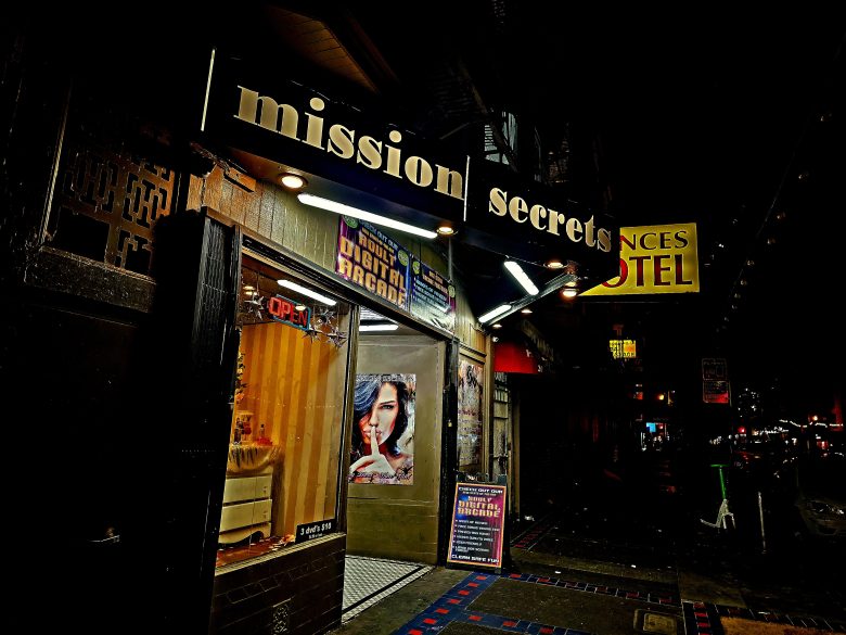 Storefront at night with a sign reading "mission Secrets," a lit "OPEN" sign, a poster of a woman, and various illuminated signs along the street.