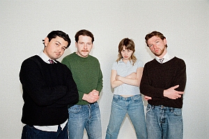 Four people stand against a plain white background, all with arms crossed and neutral expressions, wearing casual sweaters or shirts and jeans.
