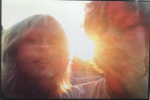 Two people with light hair stand close together outdoors, facing the camera, with sunlight shining brightly between them, partially obscuring their faces.
