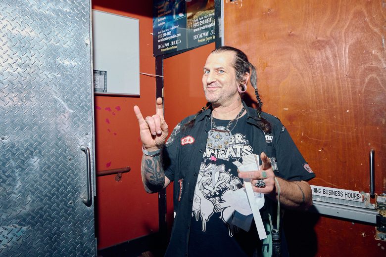 A man with tattoos and braided hair stands indoors, smiling and making the rock hand gesture. He wears a graphic t-shirt and has piercings and rings on his fingers.