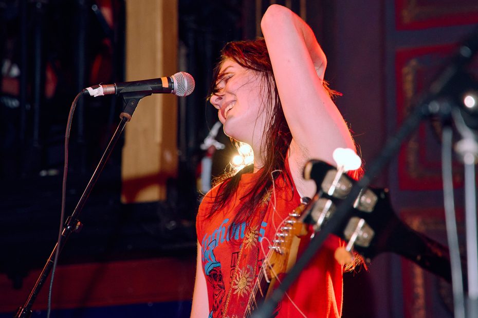 A person wearing a red shirt stands on stage, singing or speaking into a microphone, with their hand resting on their head and eyes closed.