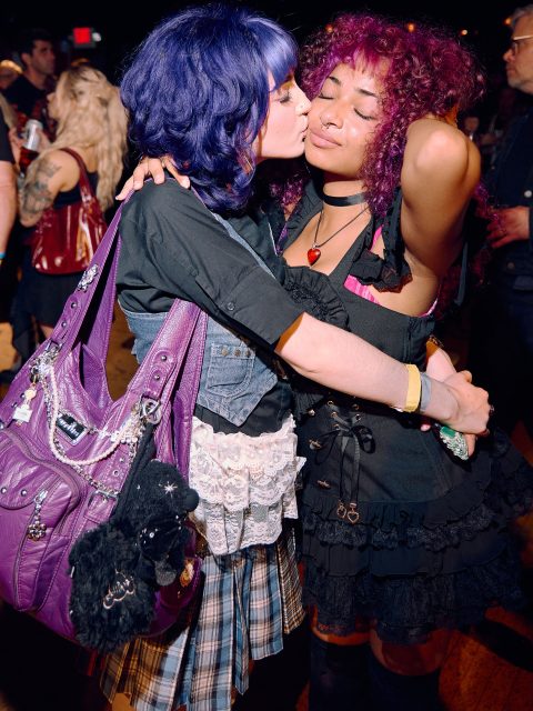Two young women with colorful hair embrace at a crowded indoor event; one kisses the other's cheek while both wear distinctive, alternative fashion.