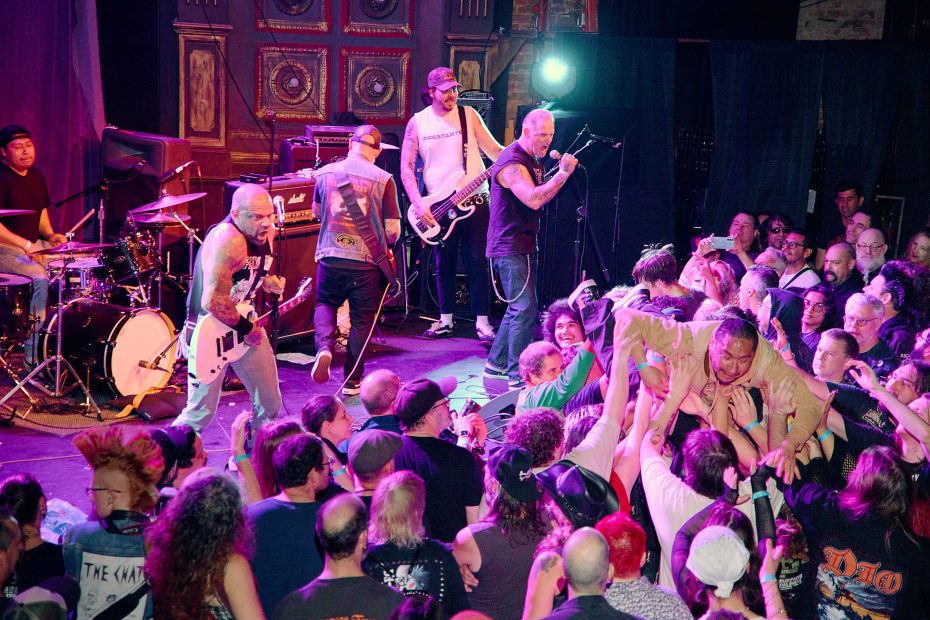 A punk band performs energetically on stage while a crowd watches and a person crowd surfs in the front.