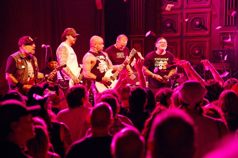 A rock band performs on stage under red lighting to a crowded audience, with multiple members playing guitars and people raising their hands.