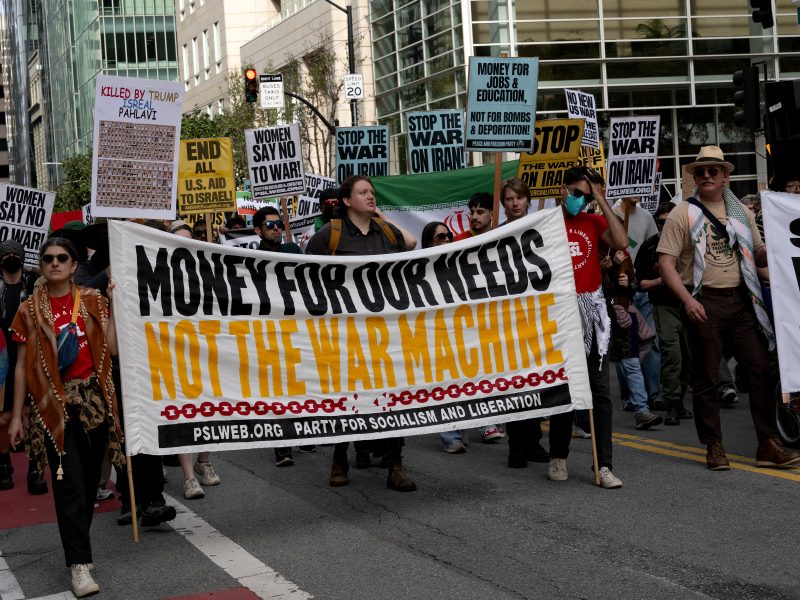 A group of protesters march on a city street holding signs, including a large banner reading "Money for our needs, not the war machine.